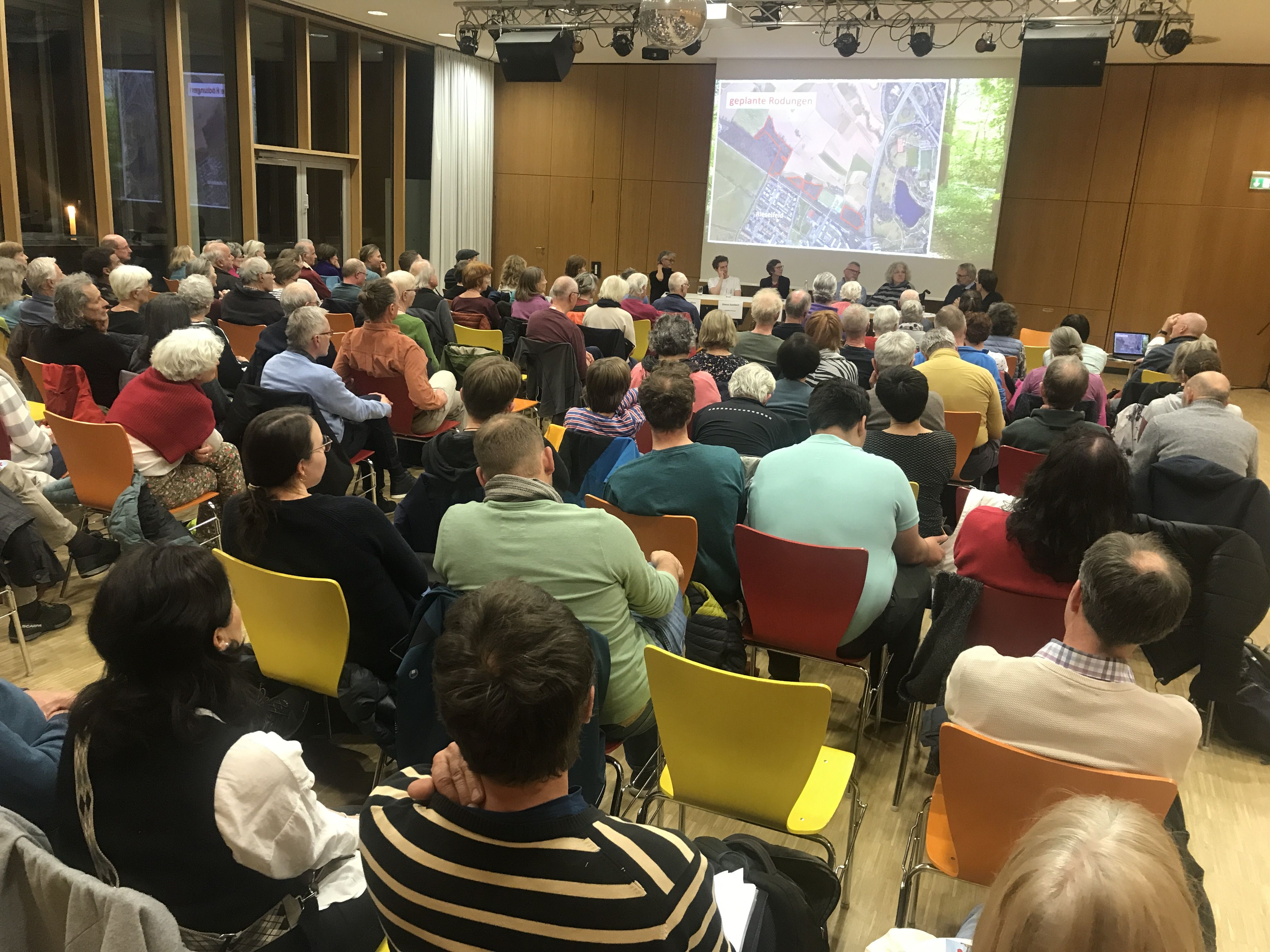 Blick auf die Rücken von Zuschauenden, die den Saal gefüllt haben auf die Leinwand an der Bühne auf der der Dietenbachwald gezeigt wird.