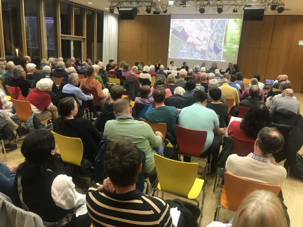Blick auf die Rücken von Zuschauenden, die den Saal gefüllt haben auf die Leinwand an der Bühne auf der der Dietenbachwald gezeigt wird.